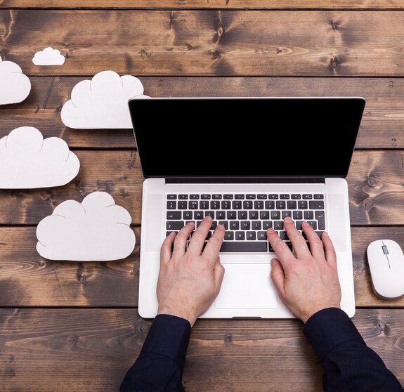 Cloud computing technology concept with white fluffy clouds next to the laptop. Mans hands typing the the keyboard uploading data, on a wooden table.