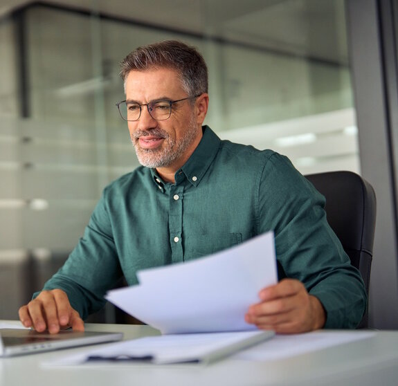 Smiling middle aged executive, mature male hr manager holding documents using laptop looking at pc computer in office at desk, checking financial data in report, doing account paper plan overview.