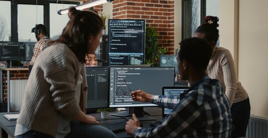 Programer sitting on desk discussing with mixed team of software developers about artificial intelligence innovation. Programmers doing teamwork looking at running machine learning algorithms.