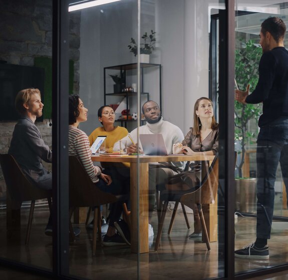 Project Manager Makes a Presentation for a Young Diverse Creative Team in Meeting Room in an Agency. Colleagues Sit Behind Conference Table and Discuss Business Development, User Interface and Design. 