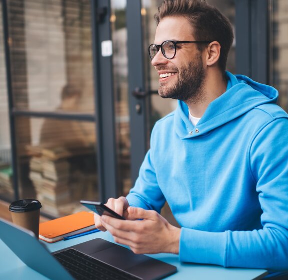 Portrait of cheerful millennial blogger with modern cellphone and laptop technology enjoying freelance lifestyle, happy hipster guy in optical eyewear using mobile phone and netbook in street cafe