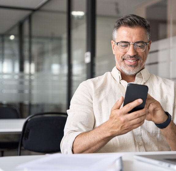 Middle aged Hispanic business manager ceo using cell phone mobile app, laptop. Smiling Latin or Indian mature man businessman holding smartphone sit in office working online on gadget with copy space.