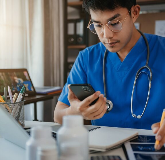 Mid adult male doctor reviews patient records on smartphone and desktop PC.