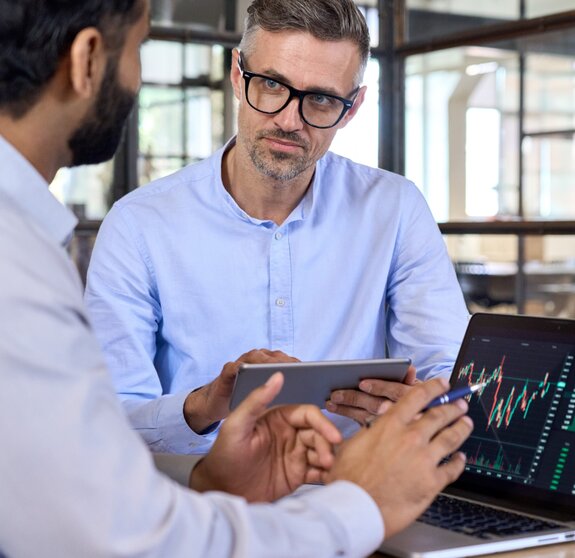 Diverse managers traders analysts discussing financial growth market at desk with laptop with graphs on screen using tablet device. Investors brokers analysing indexes online cryptocurrency stock