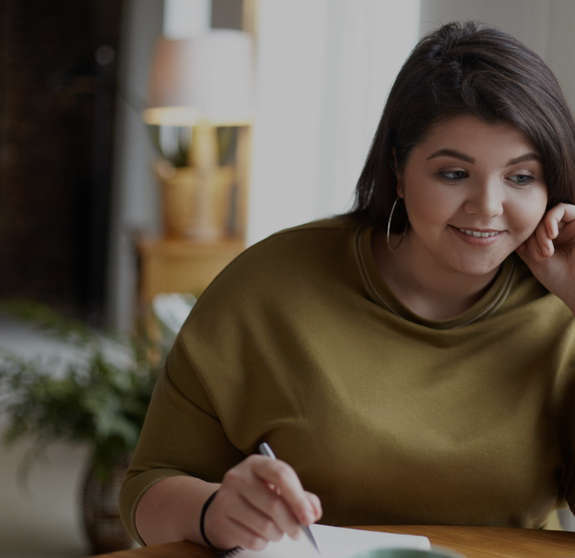 Modern electronic gadgets, job and occupation concept. Cute elegant young brunette female with excess weight using laptop computer for remote work, looking at screen, holding pen, writing down