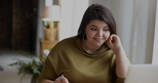 Modern electronic gadgets, job and occupation concept. Cute elegant young brunette female with excess weight using laptop computer for remote work, looking at screen, holding pen, writing down