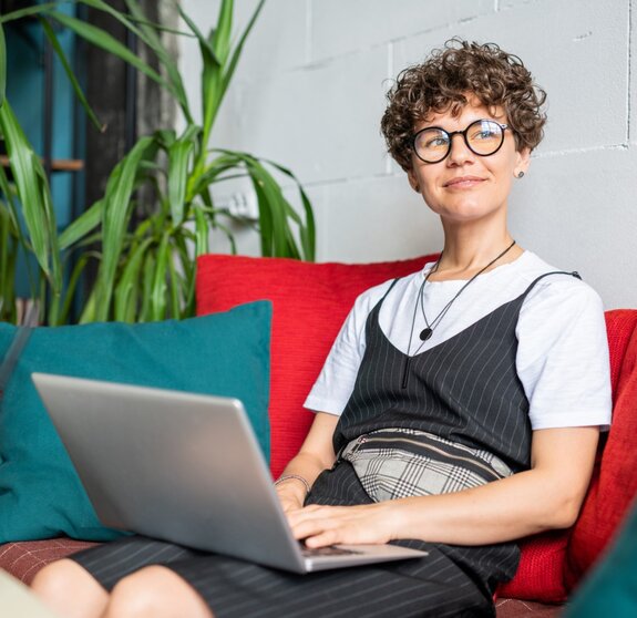 Happy young female in elegant casualwear sitting on couch with laptop in front