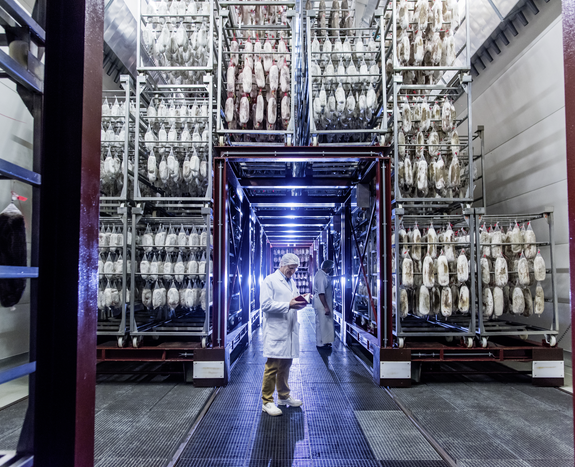 Foto einer Fabrikhalle der Bell Food Group mit zwei Mitarbeitern in Schutzkleidung und Waren die von Regalen hängen