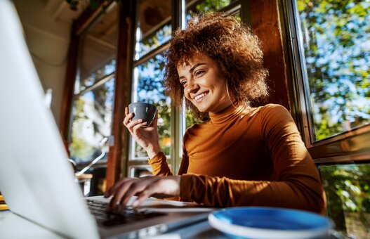 Charming young mixed race female freelancer with curly hair sitting in cafe, typing on laptop and holding cup of fresh coffee.